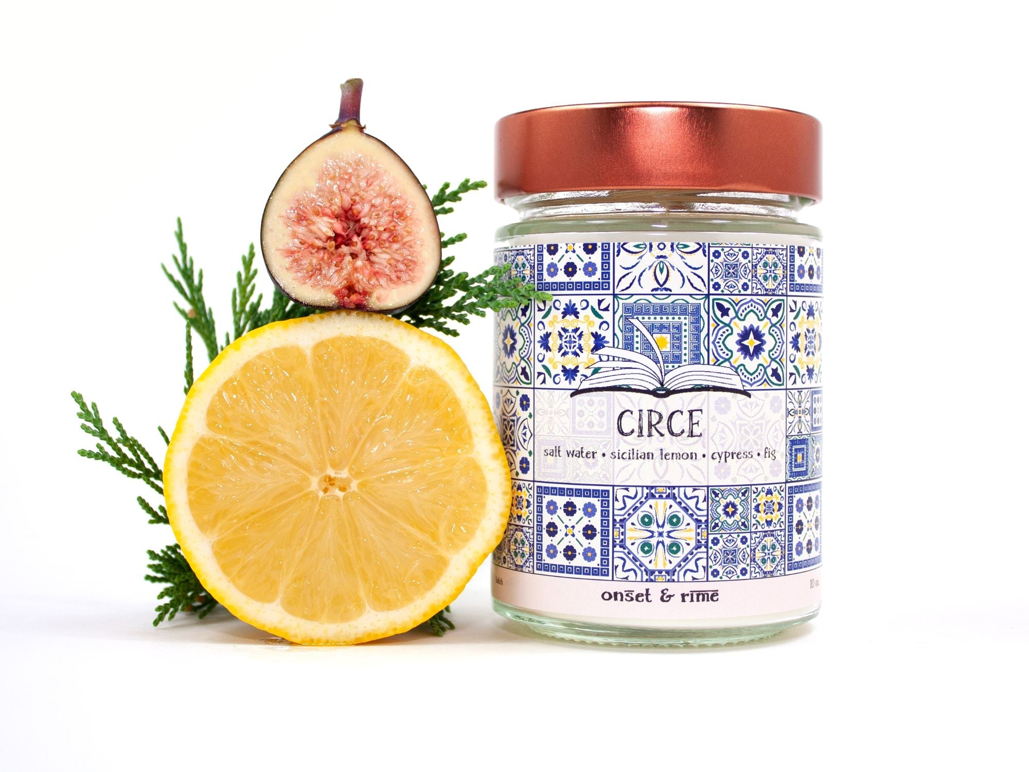 A staged photo of Onset & Rime lemon, cypress scented candle called "Circe". The candle stands next to a halved fig, a halved lemon and a branch of cypress in front of a white background.