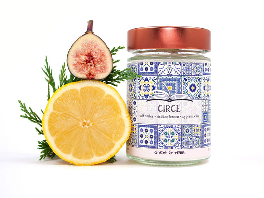 A staged photo of Onset & Rime lemon, cypress scented candle called "Circe". The candle stands next to a halved fig, a halved lemon and a branch of cypress in front of a white background.
